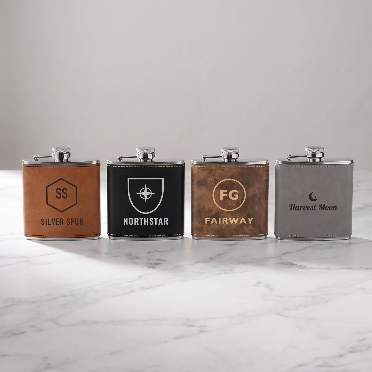 Custom logo 6 oz leatherette flasks in Rawhide, Black/Silver, Rustic/Gold and Gray, each engraved with a centered sample logo, standing on white marble