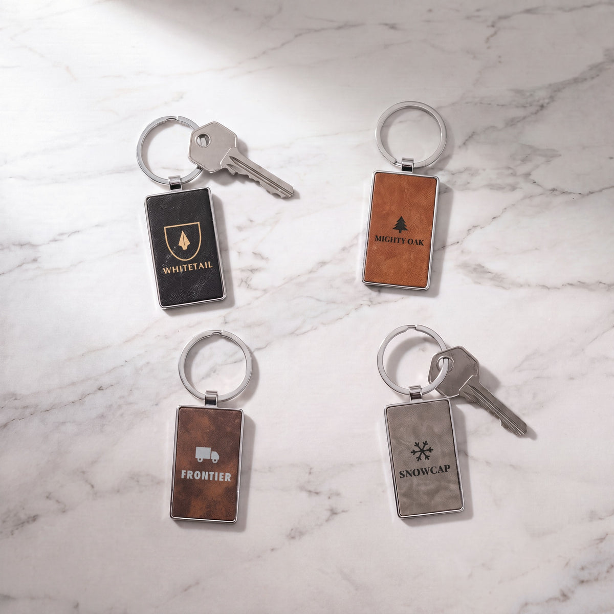 Custom Logo Leatherette & Metal Rectangle Keychains – black/gold, rawhide, rustic/silver and gray keychains on marble with split rings and keys, each engraved with a different sample logo