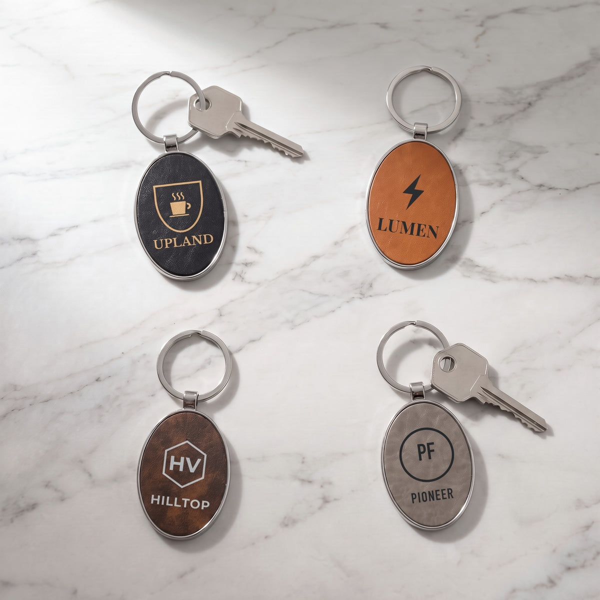 Custom logo leatherette & metal oval keychains in Black/Gold, Rawhide, Rustic/Silver and Gray, each engraved with a sample logo, on white marble