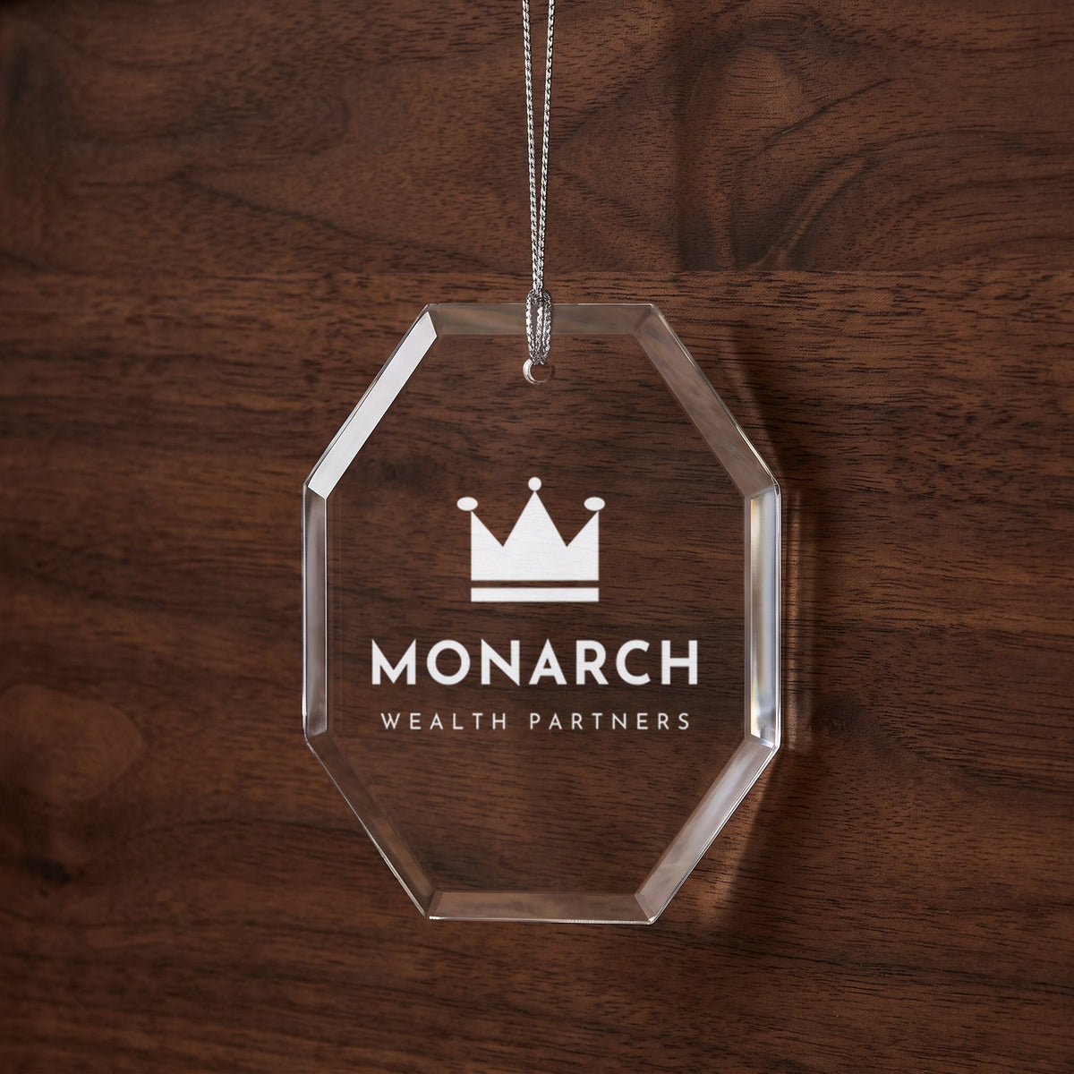 Custom Logo Glass Christmas Ornament, octagon shape with an engraved sample wealth management logo on a dark walnut wall