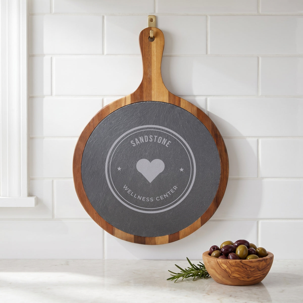 Custom Logo Round Slate & Acacia Serving Board with Handle, engraved sample logo, hanging by its handle in a kitchen